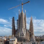Una gran basílica en construcción con varias agujas altas y una grúa en lo alto, sobre un cielo azul claro.