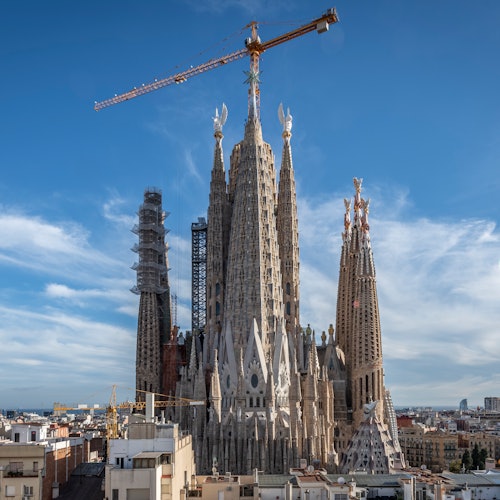 Una gran basílica en construcción con varias agujas altas y una grúa en lo alto, sobre un cielo azul claro.