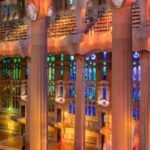 Altas columnas y coloridas vidrieras en el interior de un gran edificio ornamentado con balcones iluminados con luces cálidas y frías.