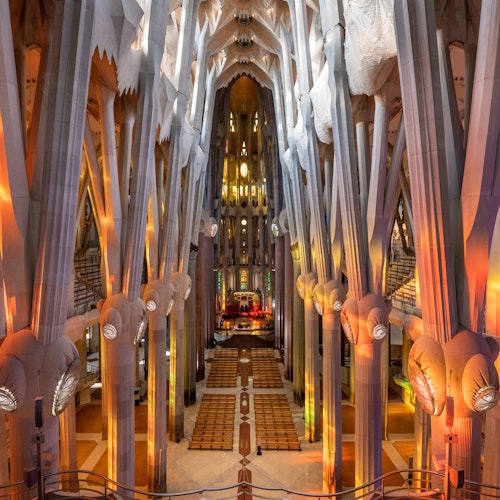 Interior de una gran catedral con altas columnas ornamentadas, vibrante iluminación de vidrieras y disposición simétrica de los asientos.