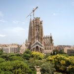 Vista aérea de la basílica de la Sagrada Familia de Barcelona rodeada de árboles y edificios de la ciudad, con una grúa visible en la parte superior.