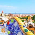 Banco de mosaico de colores con vistas a un paisaje urbano con edificios arquitectónicos únicos y un horizonte lejano bajo un cielo azul despejado.