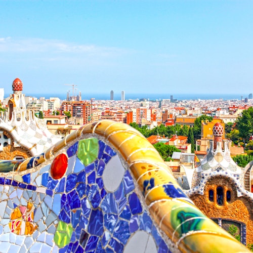 Banco de mosaico de colores con vistas a un paisaje urbano con edificios arquitectónicos únicos y un horizonte lejano bajo un cielo azul despejado.
