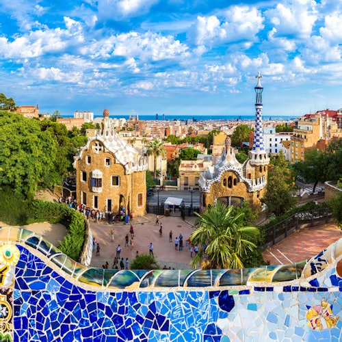 Primer plano de colorido mosaico de azulejos, gente caminando por debajo, singulares edificios con aspecto de pan de jengibre y un paisaje urbano bajo un cielo parcialmente nublado.