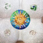 Mosaico de techo con un colorido motivo circular de rayos de sol y diseños en espiral sobre un fondo de azulejos blancos, flanqueado por detalles arquitectónicos.
