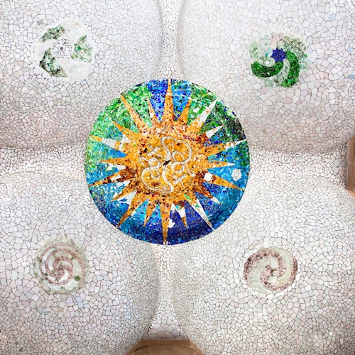 Mosaico de techo con un colorido motivo circular de rayos de sol y diseños en espiral sobre un fondo de azulejos blancos, flanqueado por detalles arquitectónicos.