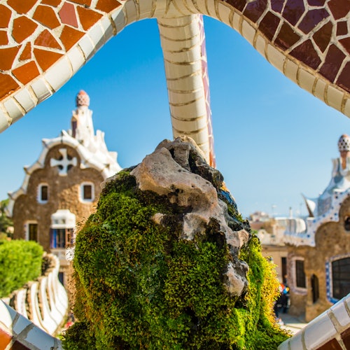Un arco de mosaico de piedra enmarca caprichosos edificios con detalles arquitectónicos únicos, vegetación y un cielo azul despejado de fondo.