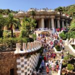 Una multitud bulliciosa explora el Park Güell de Gaudí, con su arquitectura emblemática, su exuberante vegetación y un cielo azul soleado.