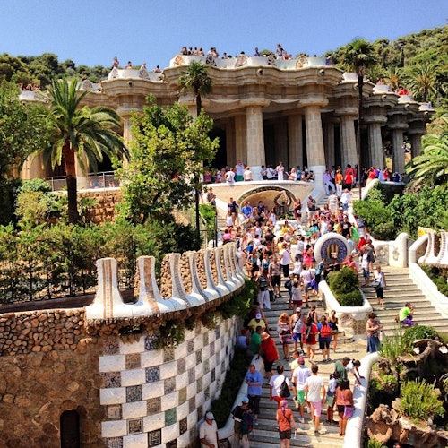 Una multitud bulliciosa explora el Park Güell de Gaudí, con su arquitectura emblemática, su exuberante vegetación y un cielo azul soleado.