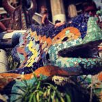 Colorida escultura de lagarto en mosaico con intrincados dibujos de azulejos, rodeada de personas y plantas en un entorno al aire libre.