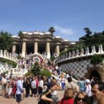 Una multitud de personas a la entrada de un parque, con una gran escalinata y columnas arquitectónicas, rodeada de vegetación.