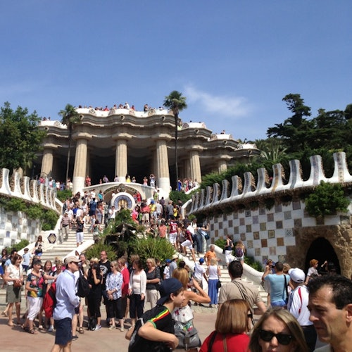 Una multitud de personas a la entrada de un parque, con una gran escalinata y columnas arquitectónicas, rodeada de vegetación.