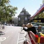 Una persona hace fotos desde el piso superior de un autobús turístico rojo, con un arco histórico y una escena de la ciudad al fondo.