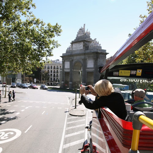 Una persona hace fotos desde el piso superior de un autobús turístico rojo, con un arco histórico y una escena de la ciudad al fondo.