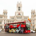 Un autobús rojo de dos pisos con el rótulo "Madrid City Tour" delante de un ornamentado edificio blanco con banderas españolas.
