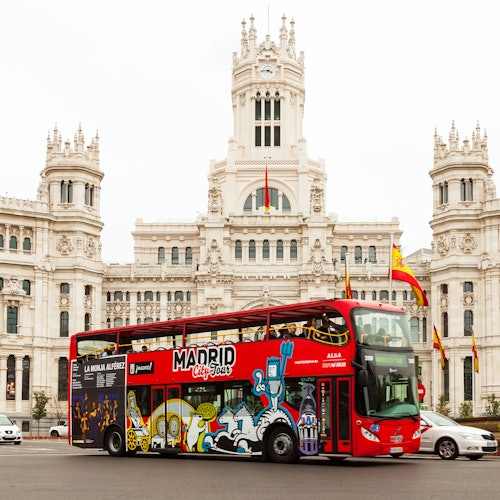 Un autobús rojo de dos pisos con el rótulo "Madrid City Tour" delante de un ornamentado edificio blanco con banderas españolas.