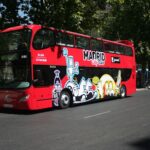 Autobús rojo de dos pisos con la marca "Madrid City Tour" e ilustraciones de dibujos animados de colores en el lateral, aparcado en una calle.