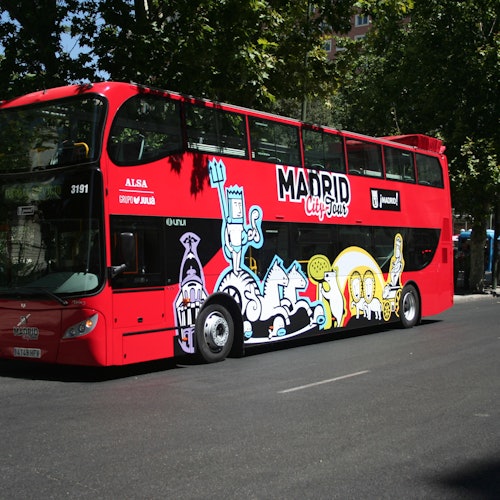 Autobús rojo de dos pisos con la marca "Madrid City Tour" e ilustraciones de dibujos animados de colores en el lateral, aparcado en una calle.