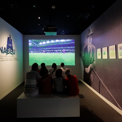 A group of people watching a soccer match on a screen in a dim room with images and text on the side walls.