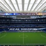 Interior view of a large stadium with green field, blue seating, and "REAL MADRID" displayed in white letters on the stands.