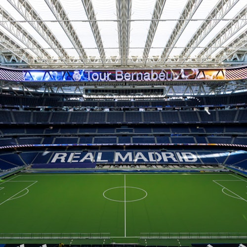 Interior view of a large stadium with green field, blue seating, and "REAL MADRID" displayed in white letters on the stands.