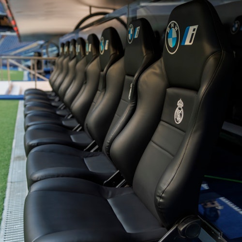 A row of black leather stadium seats with BMW and Real Madrid logos, positioned on a sports field's sideline.