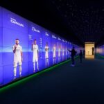 A futuristic display wall showing life-sized images of soccer players in a dimly lit room with visitors interacting.