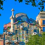Un edificio colorido y ornamentado, con tejado ondulado, tejas decorativas y formas singulares, enmarcado por verdes árboles bajo un cielo azul.