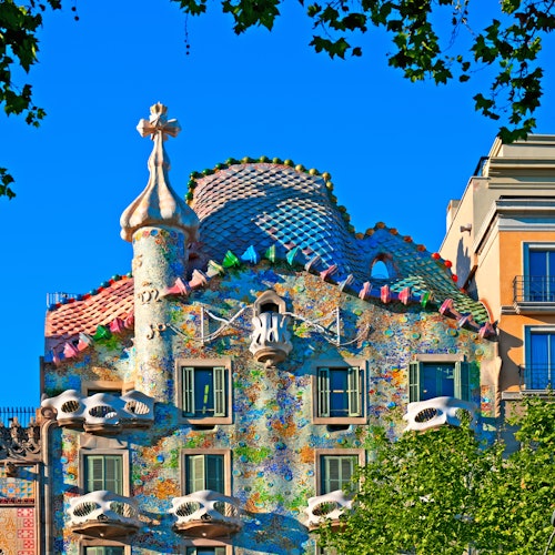 Un edificio colorido y ornamentado, con tejado ondulado, tejas decorativas y formas singulares, enmarcado por verdes árboles bajo un cielo azul.