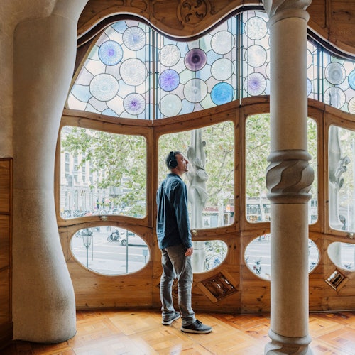 Casa Batlló: Entrada Una persona con auriculares está de pie dentro de una habitación con grandes vidrieras de colores y marcos de madera ornamentados.