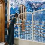Una mujer con un vestido largo escucha unos auriculares mientras mira por encima de una barandilla de cristal ondulado y reflectante en un espacio embaldosado de tonos azules.