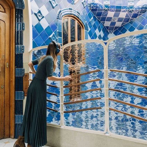 Una mujer con un vestido largo escucha unos auriculares mientras mira por encima de una barandilla de cristal ondulado y reflectante en un espacio embaldosado de tonos azules.